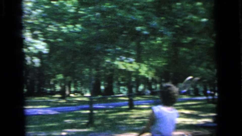 A woman in a white top is seen from behind as she swings or lifts a badminton racket in the Camden park scene.