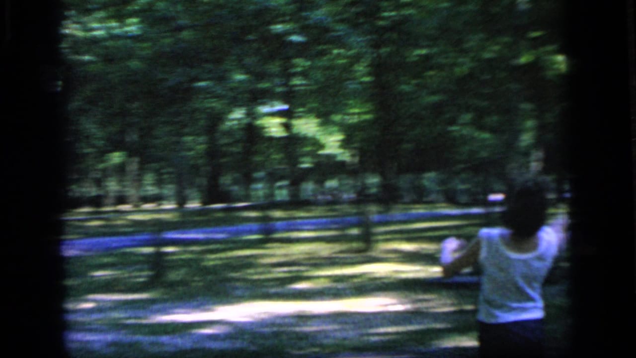 Woman in a white sleeveless top stands in a wooded park during a badminton game.