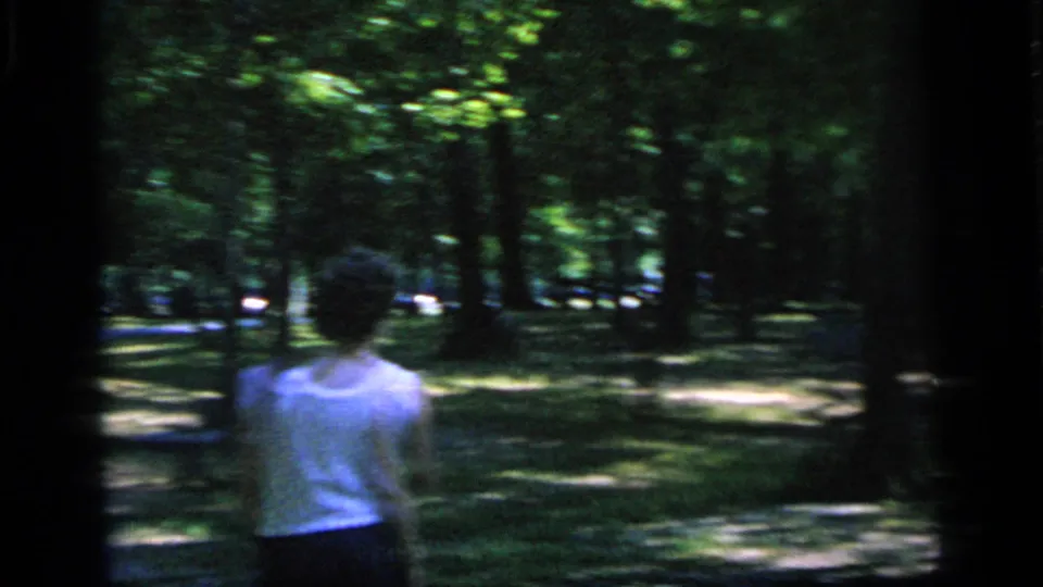 A soft 8mm frame from Camden in 1964 shows a woman from behind on grass near trees during the badminton outing.