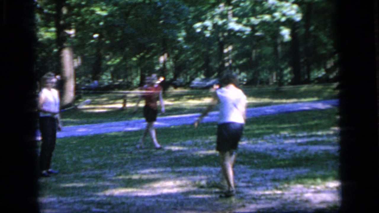In metadata-supported 1964 Camden, three adults appear to play casual tennis on grass beside a shaded park path.