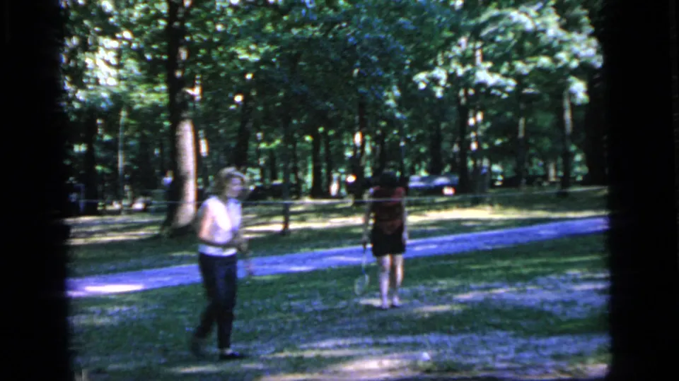 Two casually dressed adults with rackets stand in the Camden park, framed by trees, grass, and a paved path.