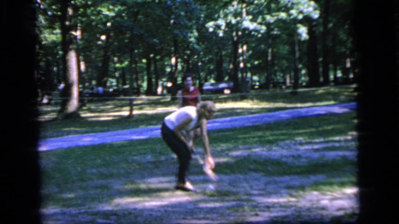 In Camden, New Jersey in 1964, a woman in a white blouse bends on grass near a path while another adult stands behind her.