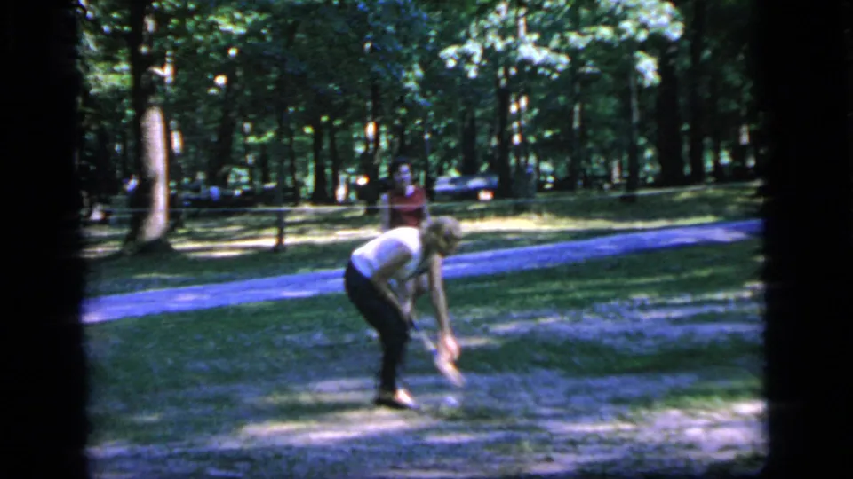 In Camden, New Jersey in 1964, a woman in a white blouse bends on grass near a path while another adult stands behind her.