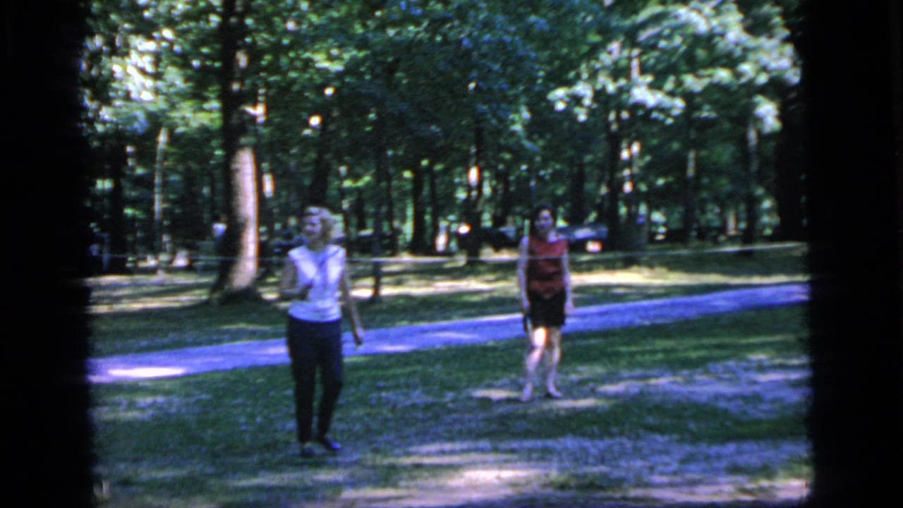 Two adults stand near a paved path in a wooded park.