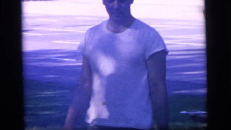A man in a white T-shirt is seen in soft focus near a road or parking area during the 1964 Camden home-movie scene.