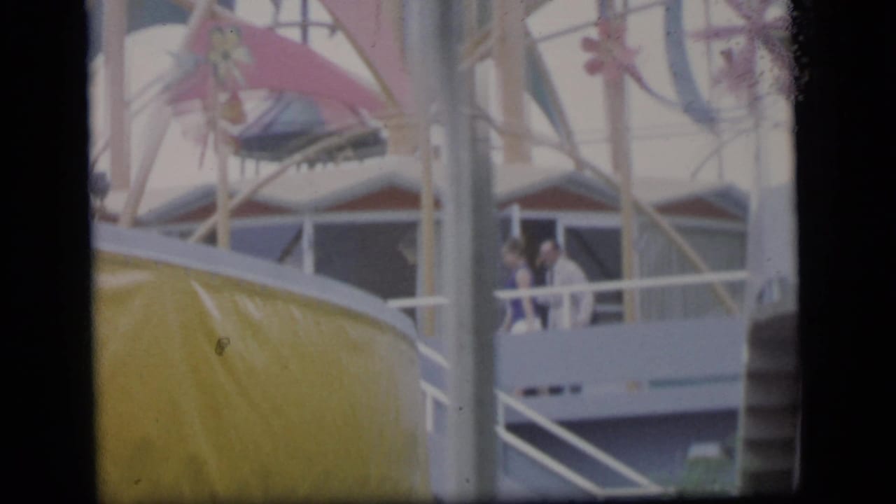 Camden 1964 home-movie frame showing distant people on a balcony near railings and a yellow curved structure.