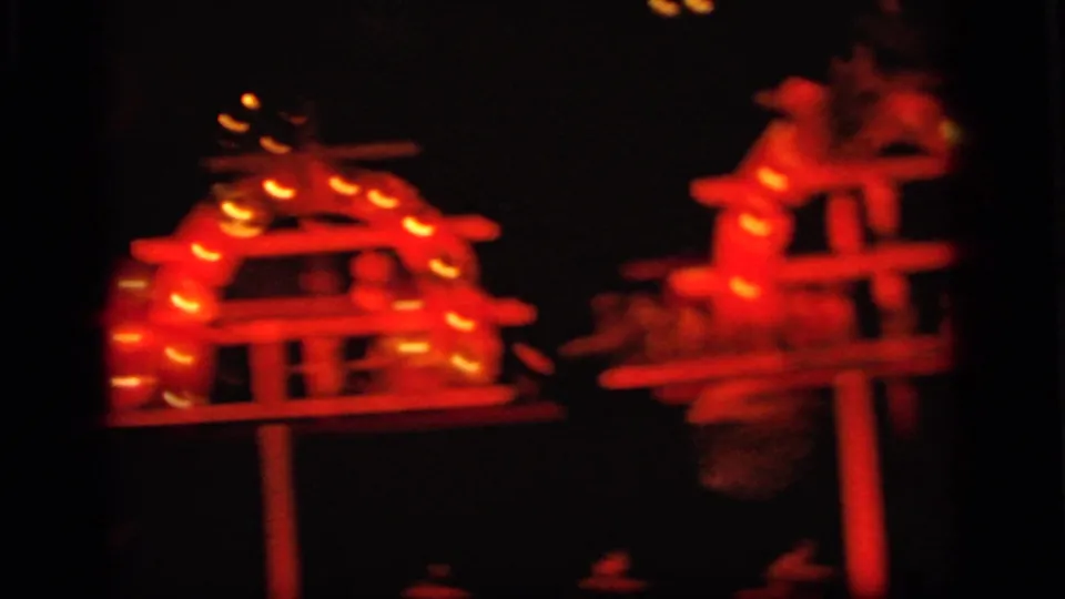 Red-lit 1964 8mm frame showing illuminated puppet-like figures on small raised stage structures in Camden footage.