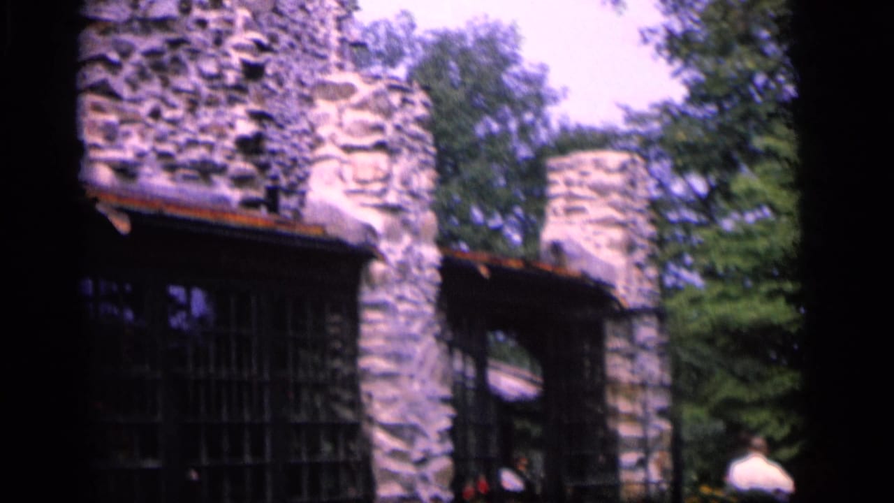 1964 Camden, New Jersey frame showing a rough stone structure with arched openings, trees, and a distant visitor at the edge.