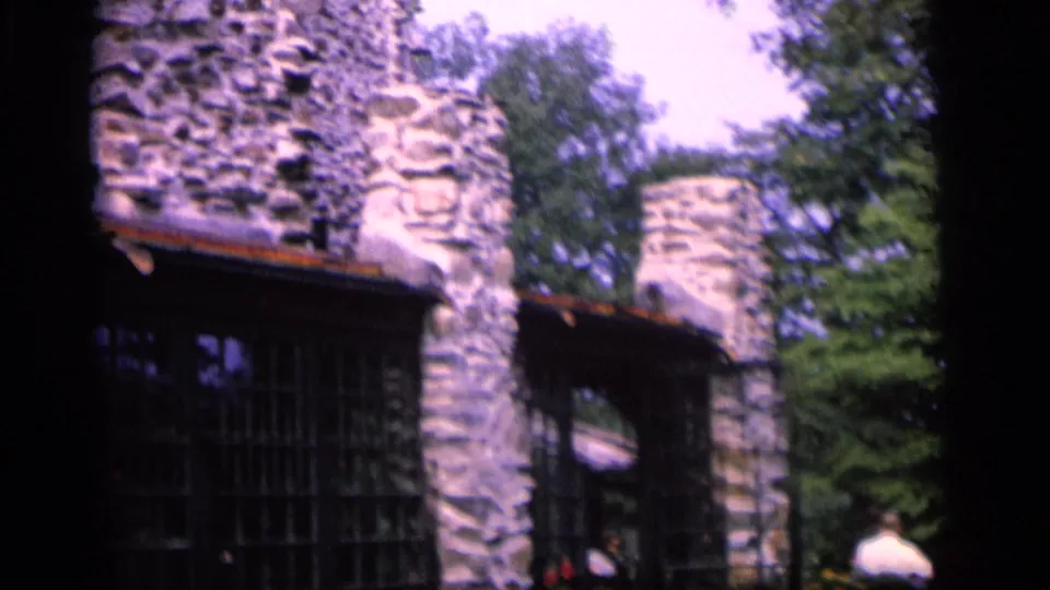 1964 Camden, New Jersey frame showing a rough stone structure with arched openings, trees, and a distant visitor at the edge.