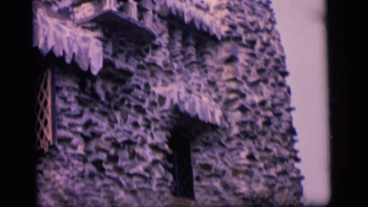 1964 Camden home-movie frame showing a rough stone facade with dark window openings and scalloped thatch-like eaves.