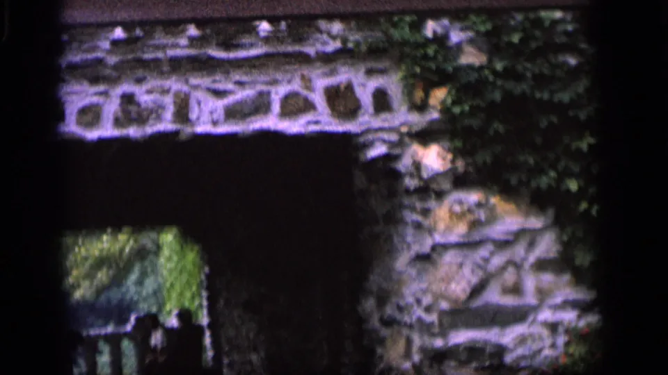 1964 Camden home-movie frame tilting down to a stone archway, ivy, railing, and small shadowed figures below.