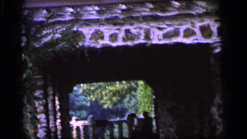 1964 Camden frame showing a shadowed stone arch, rough masonry, greenery beyond, and a small silhouetted figure.