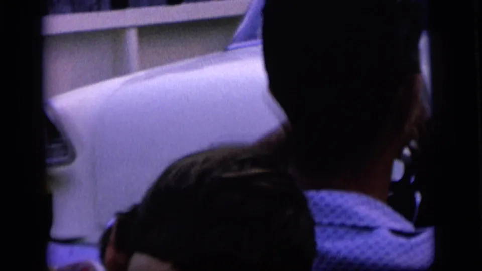 1964 home-movie frame with partial family figures near a light-colored car at the lakeside.