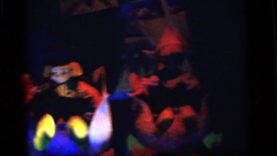 Dim 8mm frame with toy-like figures, red objects, and saturated blue light in a Camden, New Jersey home-movie setting.