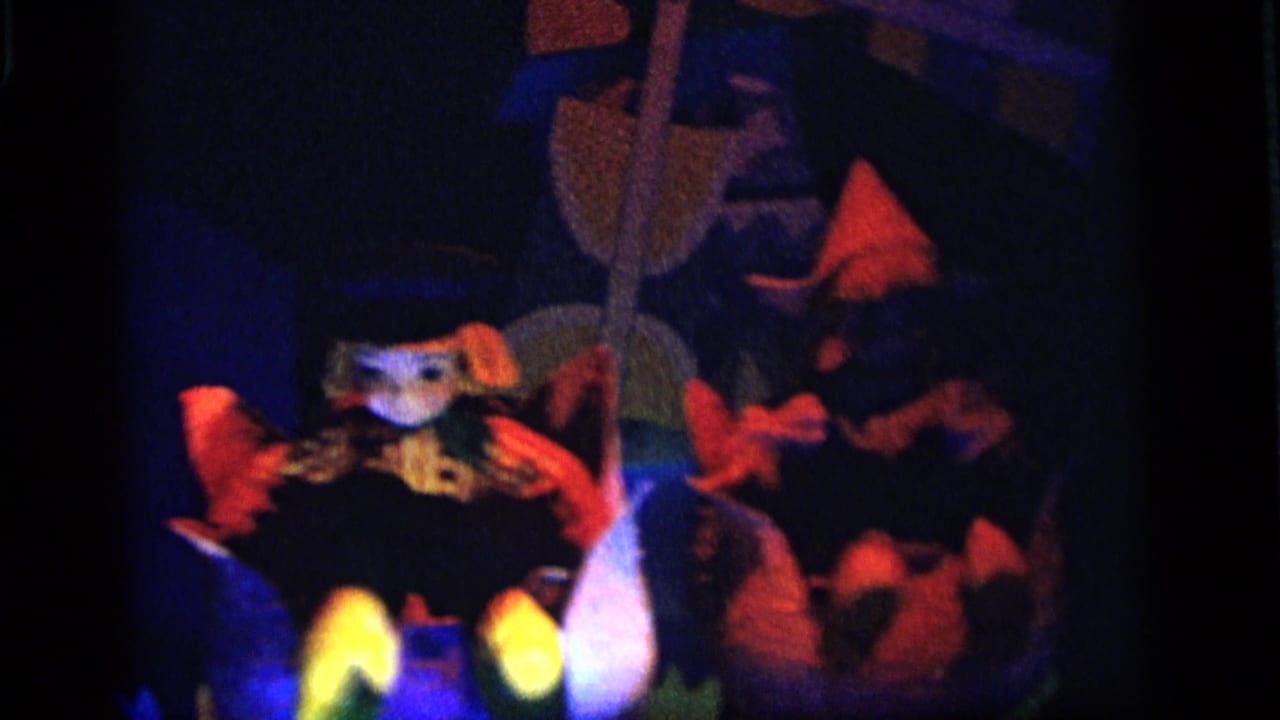 1964 Camden home-movie frame showing a dark puppet stage with a seated figure, pointed hats, red accents, and blue theatrical light.