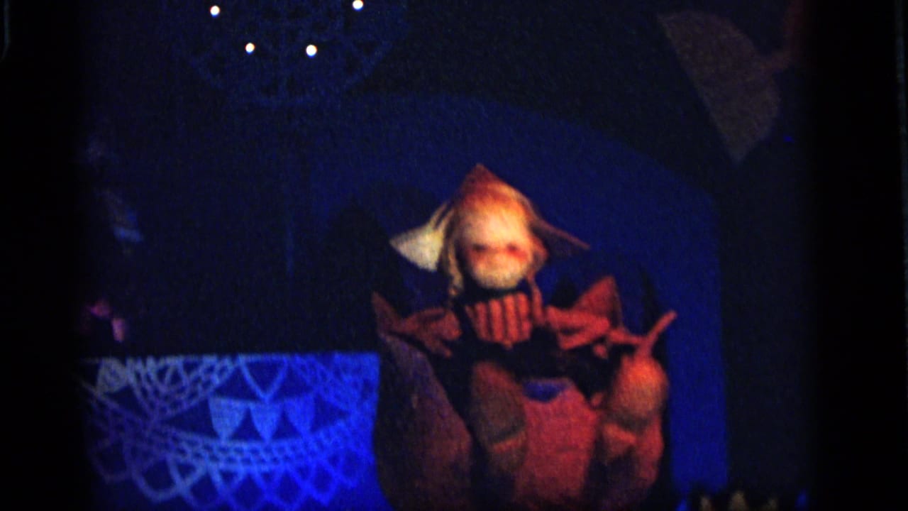 Puppet with pale hair and wide collar posed against a dark blue stage.