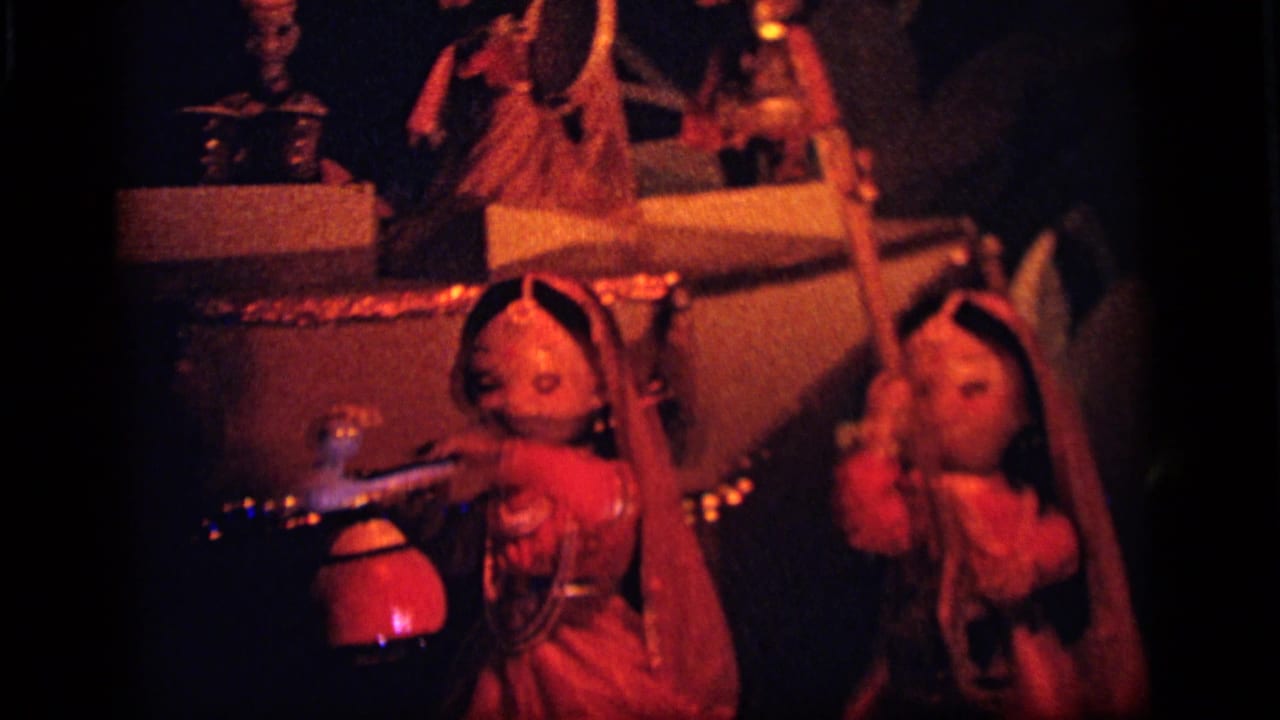 1964 8mm frame from Camden showing two veiled toy dolls posed in a red-orange illuminated display with small props and platforms.