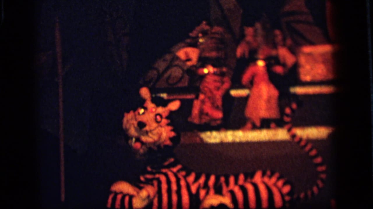 Camden 1964 home-movie frame with a striped tiger puppet in front of a dim red-orange toy performance display.