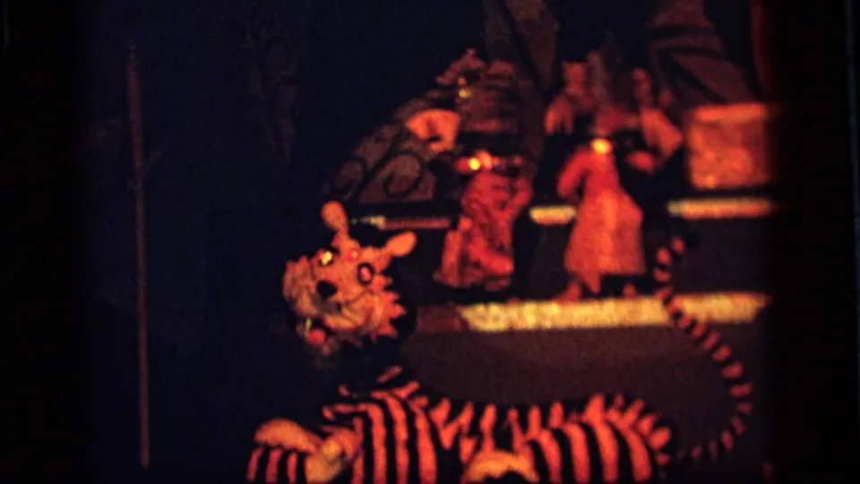 Camden 1964 home-movie frame with a striped tiger puppet in front of a dim red-orange toy performance display.