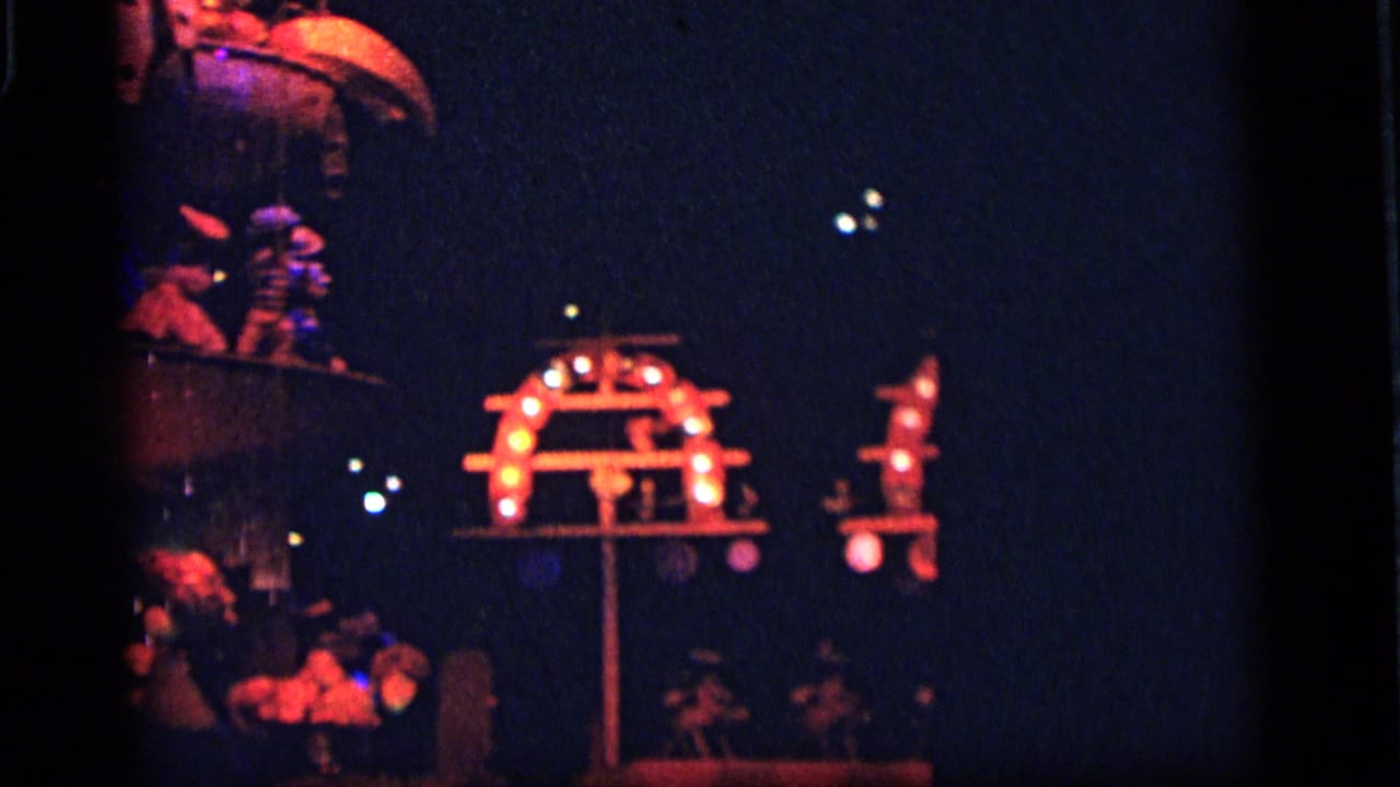 1964 Camden amusement-park frame with a small bulb-lit structure, dark surrounding space, and partial attraction figures at the edges.