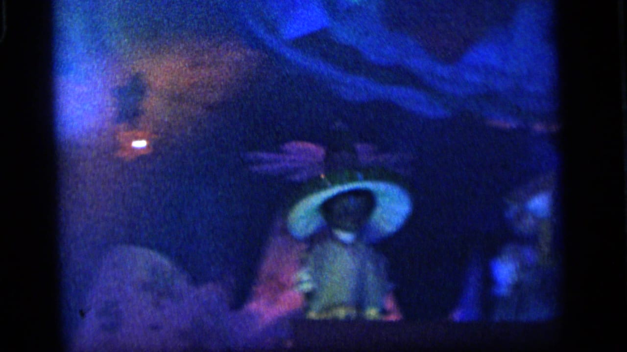 1964 Camden home-movie frame showing a soft-focused blue-lit toy stage with a small doll figure and painted backdrop shapes.