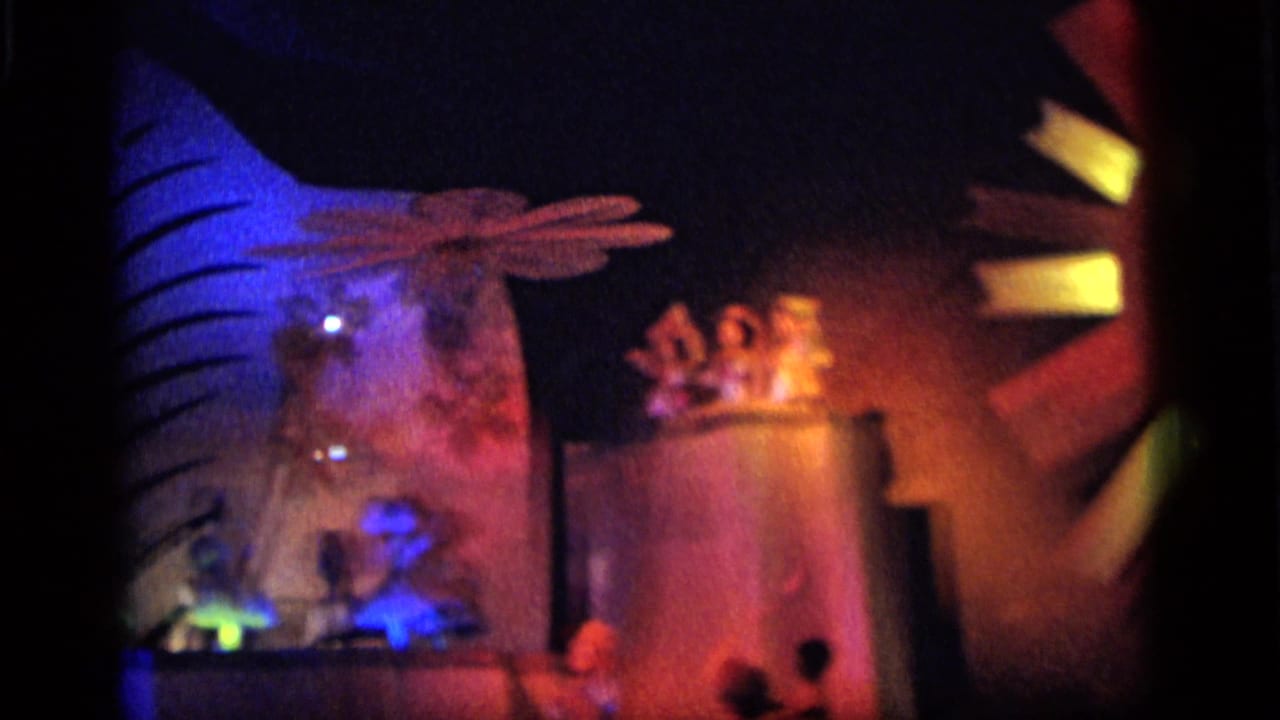 1964 Camden footage shows a dim toy theater stage with small figures, glowing set pieces, and a large sun-face element at the edge.