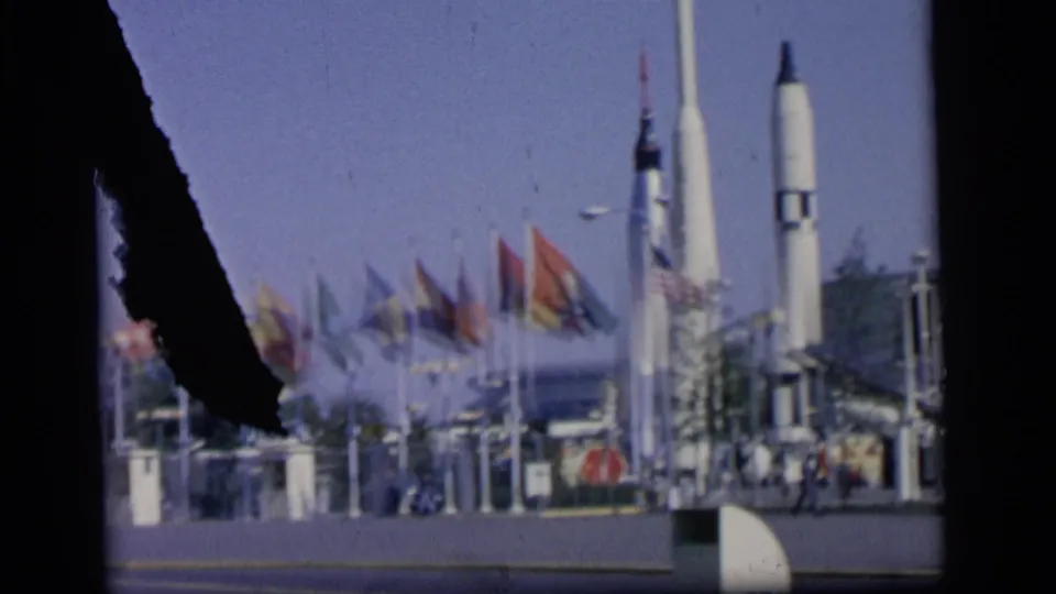 1964 Camden home-movie frame showing national flags and tall rocket displays behind a dark film-edge obstruction.