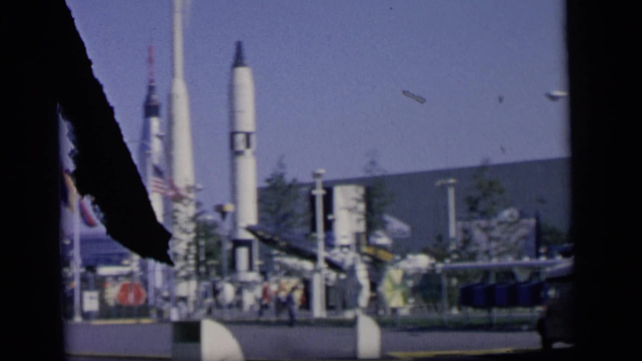 1964 Camden frame with rocket and missile displays, national flags, exhibit structures, and small visitors partly obscured by film damage.
