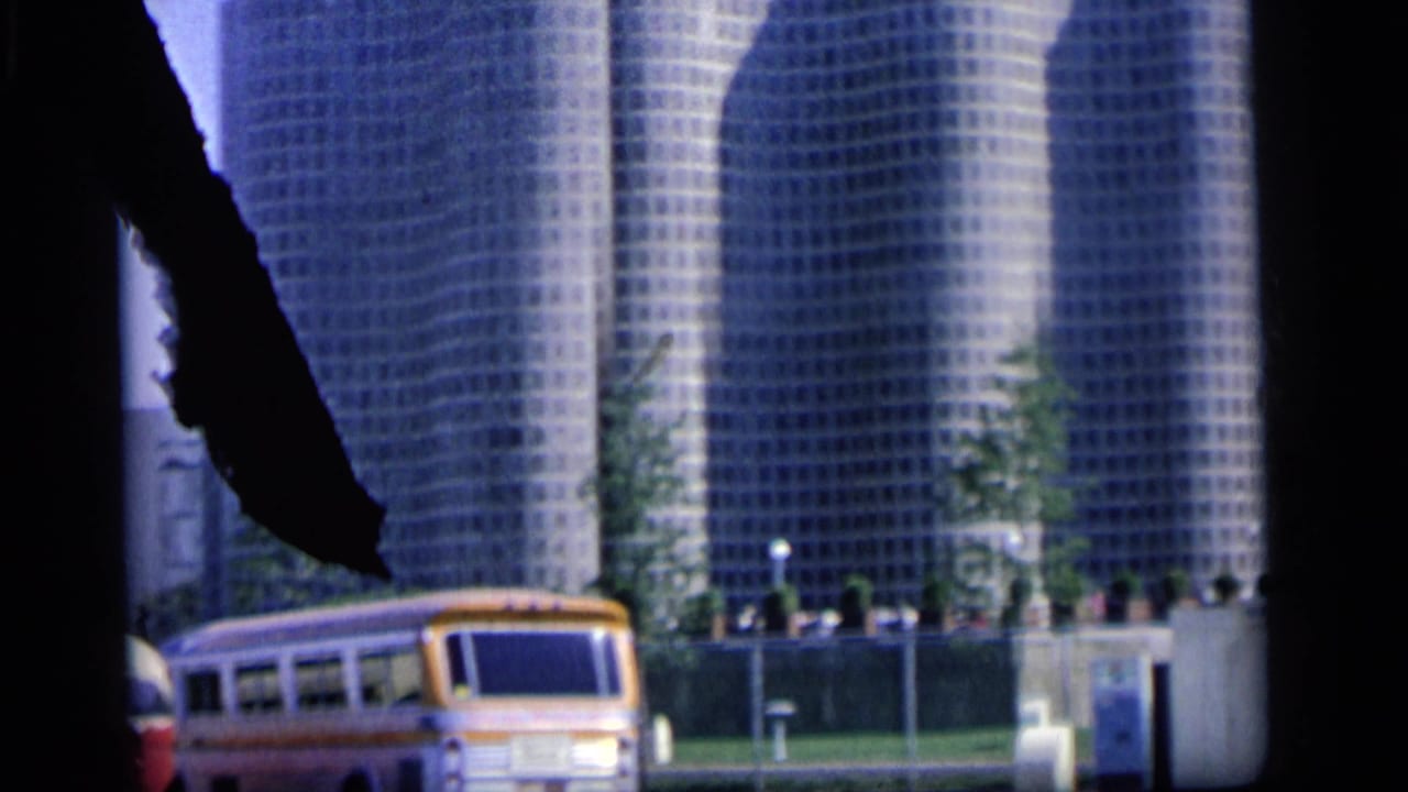 Grid-patterned office towers behind a yellow and white bus, framed by dark film or window edges.