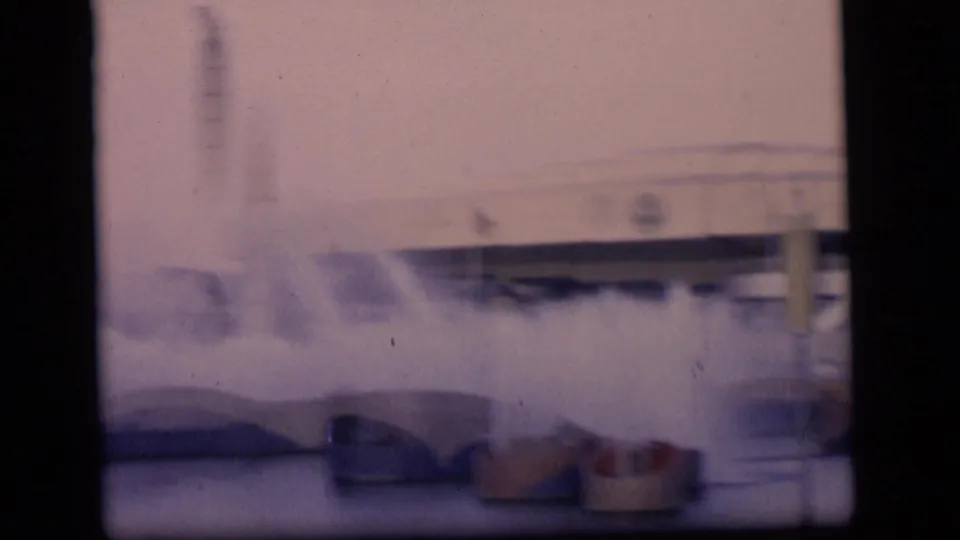 1964 Camden-area 8mm frame showing fountain sprays through a window frame, with soft buildings and a tall stack behind them.