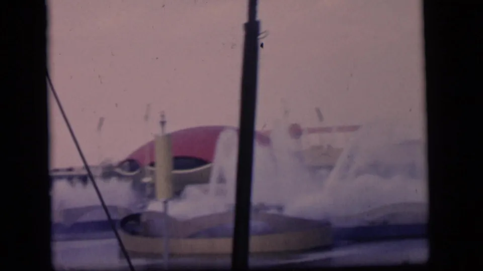 1964 Camden, New Jersey home-movie frame with water jets, a red-domed structure, a yellow pole, and dark window mullions.