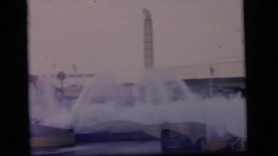 1964 Camden-area 8mm frame with heavy fountain spray, a tall smokestack-like structure, low basins, and soft background architecture.