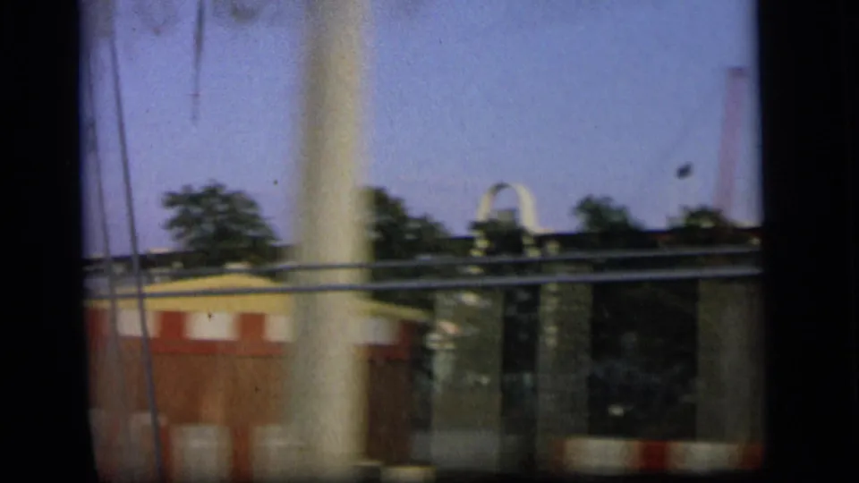 Final 1964 Camden frame with roadside railing, trees, poles, and a partial striped tent edge blurred by vehicle movement.
