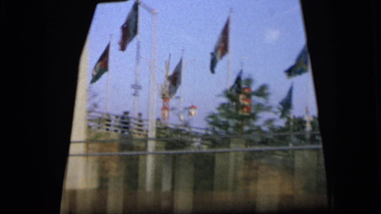 1964 Camden view from inside a moving car, with multiple flags on poles behind a roadside fence.