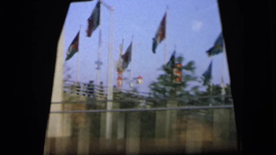 1964 Camden view from inside a moving car, with multiple flags on poles behind a roadside fence.