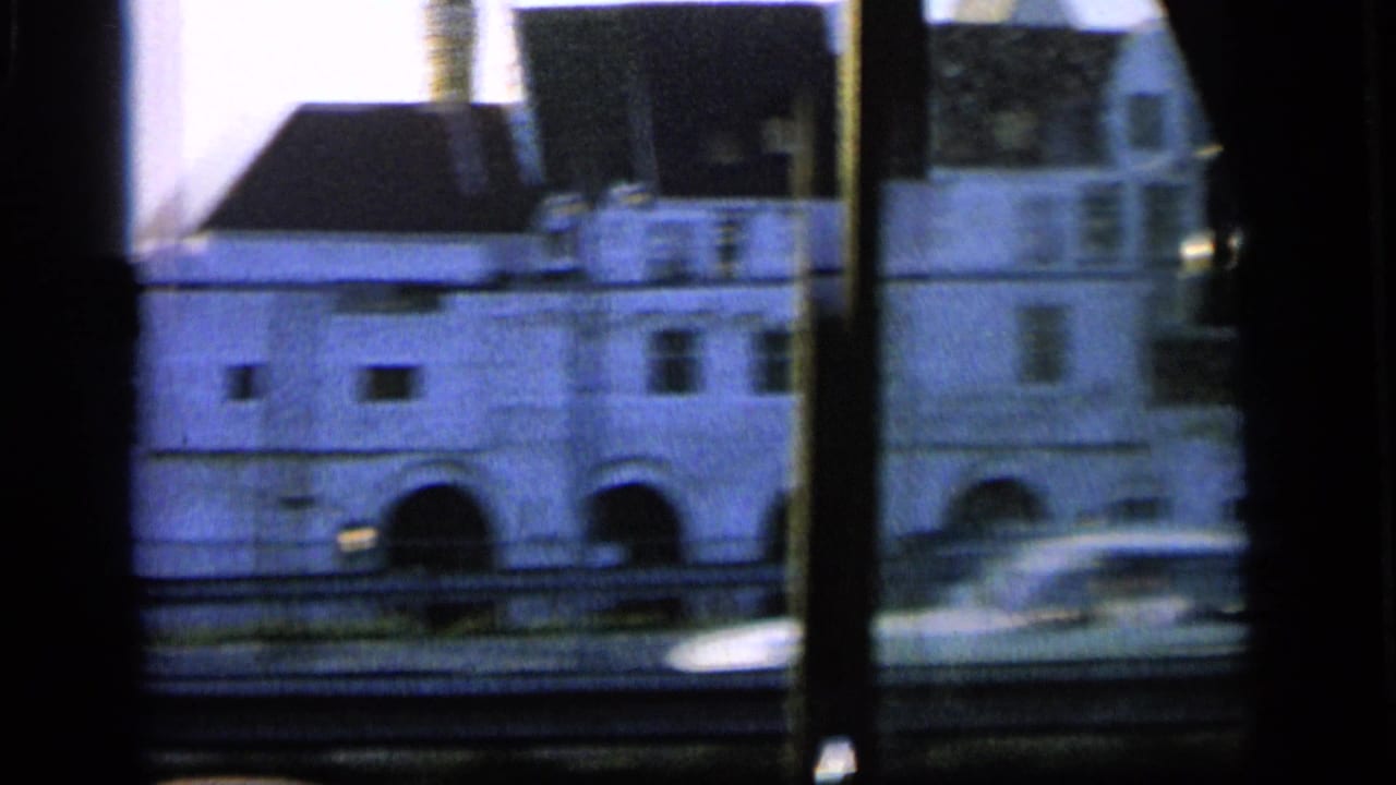 View through a vehicle window of pale buildings with arched openings and dark roofs.