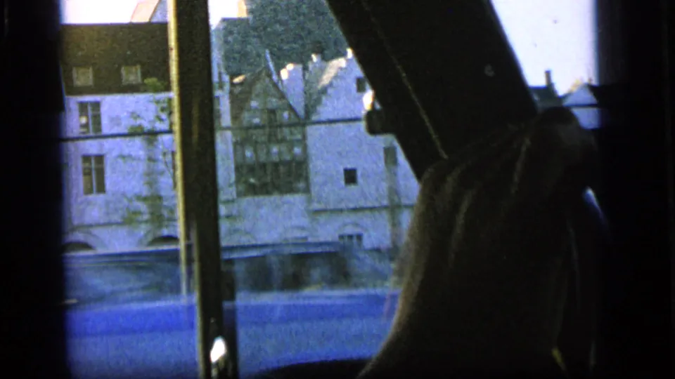 Inside a moving vehicle in Camden, a white building and roadside view pass behind window supports while a passenger's arm appears at right.