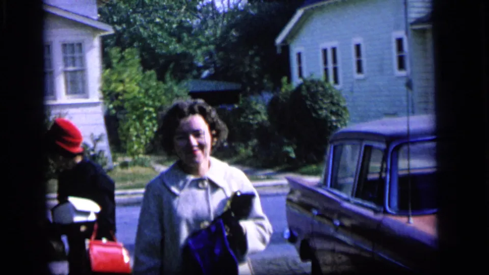 A woman in a light coat stands near a parked 1960s-era car on a residential street, with another person in a red hat at left.