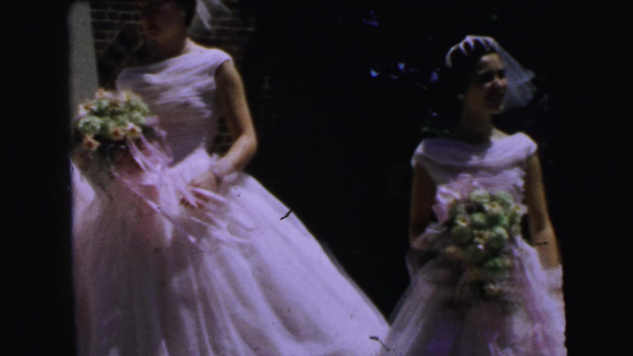 Two bridesmaids in white gowns and veils wait outside in the 1964 Camden wedding home movie, each holding a bouquet.