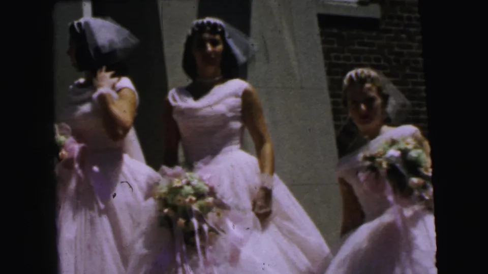 Three bridesmaids in white gowns, veils, and bouquets stand outside a brick and light-colored wall in the 1964 Camden wedding clip.