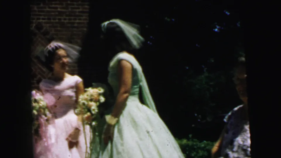 A Camden wedding home-movie frame shows two bridal party members speaking outside, with bouquets and greenery nearby.