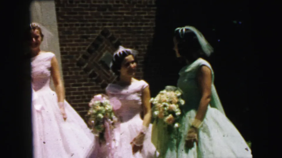 1964 Camden bridal party frame with women in pale formal dresses holding bouquets near brickwork and a shaded exterior.