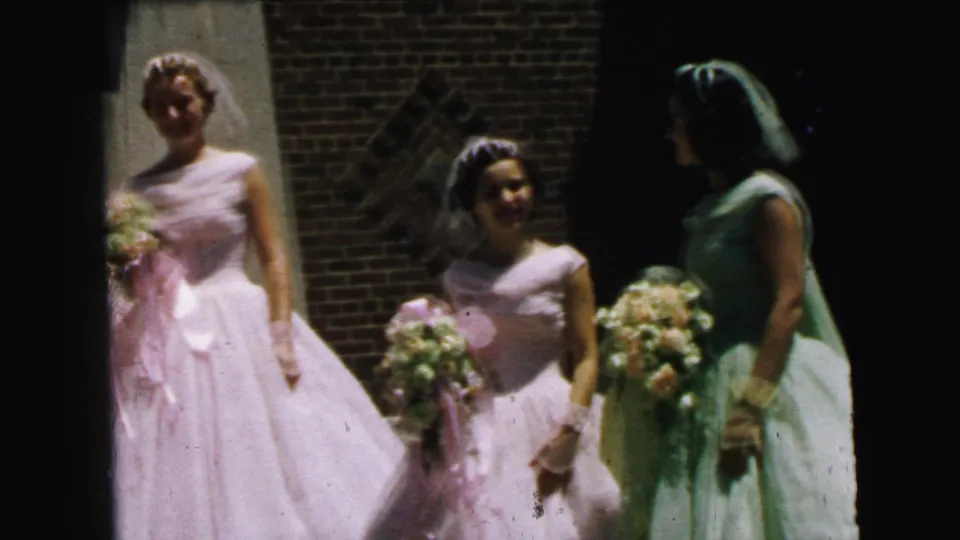 1964 Camden home-movie frame showing bridesmaids in pale pink and green dresses with veils and bouquets outside a brick entrance.