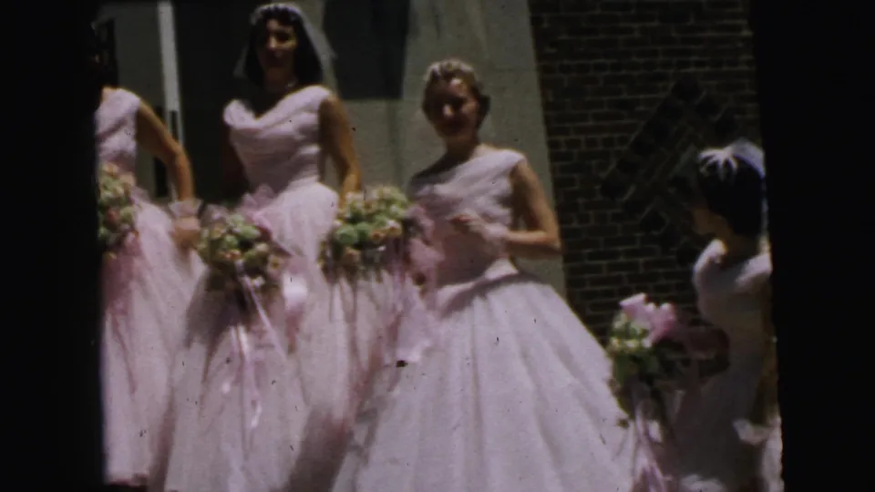 1964 Camden home-movie frame of bridesmaids grouped outside a brick building, with bouquets, veils, and pale formal gowns visible.