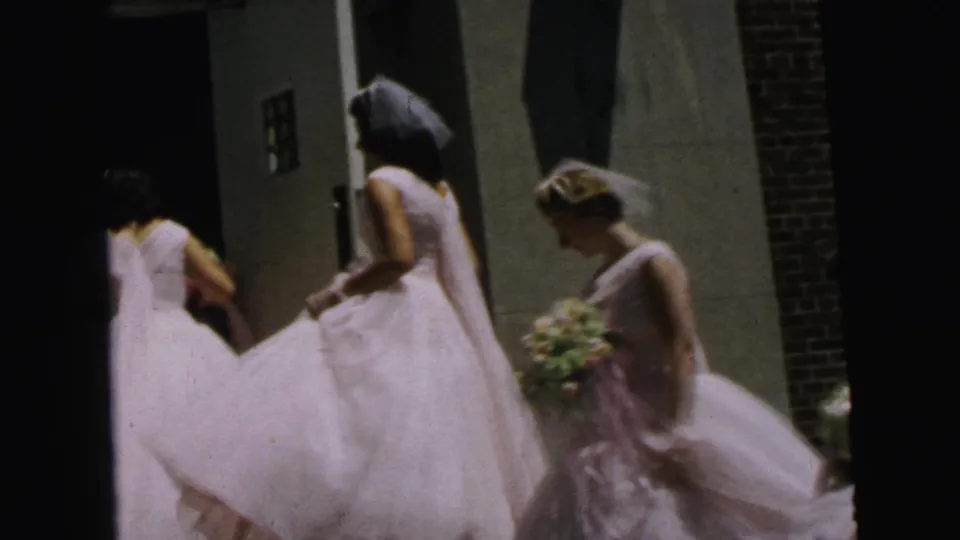 1964 Camden frame showing bridesmaids in pale dresses moving near the building entrance, with one holding a gown and another carrying flowers.