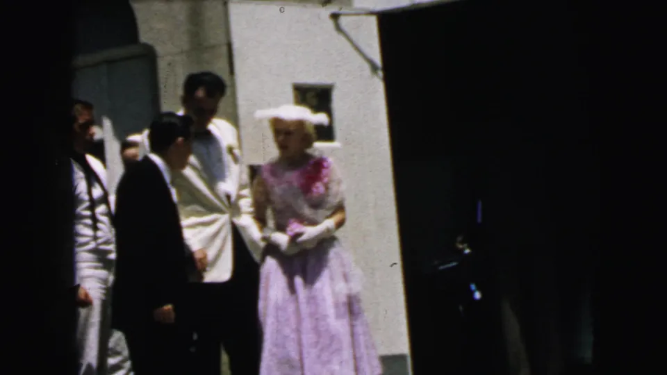 1964 Camden church exterior with a woman in a lavender dress and white hat beside men in formal wear, seen in bright sun.