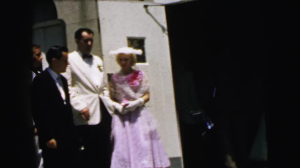 1964 Camden wedding-party frame showing a man in a white dinner jacket, a woman in lavender, and nearby formal guests outside church.