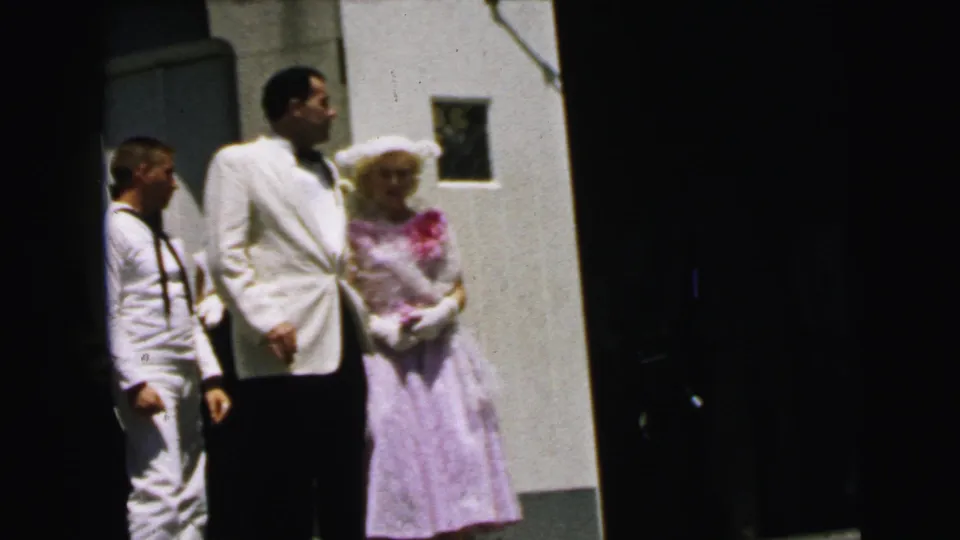 1964 Camden 8mm frame of wedding-party members outside a church, centered on a man in a white jacket and woman in lavender.