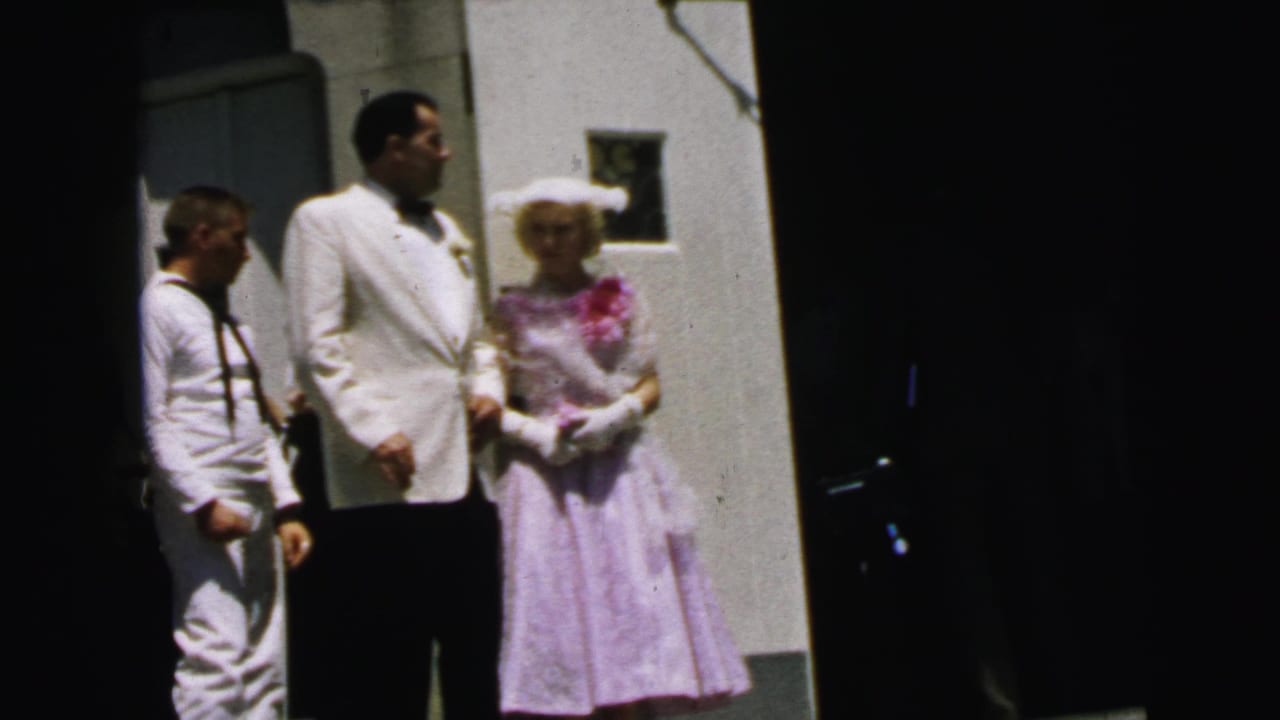 Man in a white jacket stands beside a woman in a lavender dress and a sailor in white uniform.