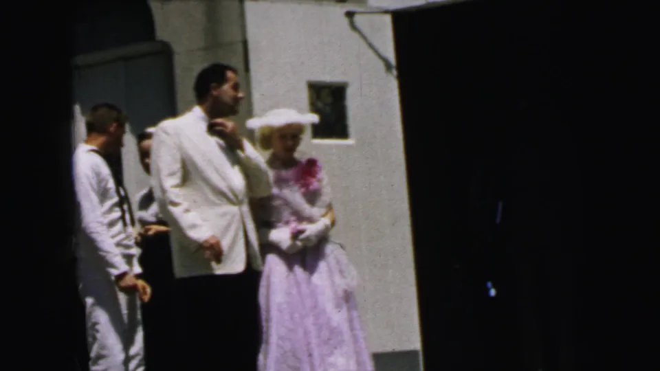 1964 Camden home-movie still showing the man touching his bow tie as the woman and sailor stand nearby.